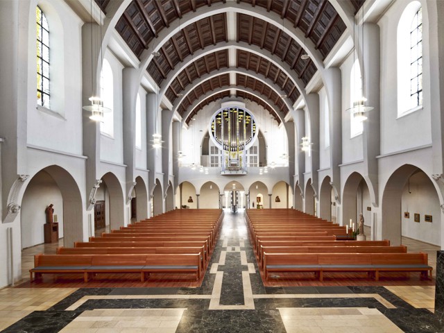 Das Bild zeigt eine Ansicht eines leeren Kircheninneren mit Bänken, die an den Seiten angeordnet sind, und einem großen Bleiglasfenster am Ende des Hauptschiffs.