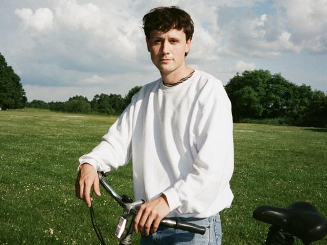Ein junger Mann, der sich mit seinem Fahrrad auf einer grünen Wiese präsentiert und lässige Kleidung sowie eine Sonnenbrille trägt