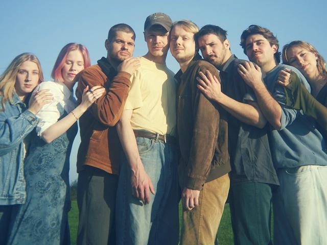 Gruppenfoto von sieben Freunden mit verschiedenen Hairstyles und Farben, die lachend vor der Kamera posieren, auf einer grasbewachsenen Wiese unter einem blauen Himmel.