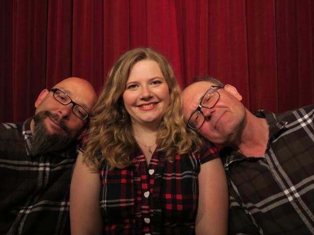 Eine junge Frau, die für ein Foto mit zwei älteren Männern in Karoschirts posiert.