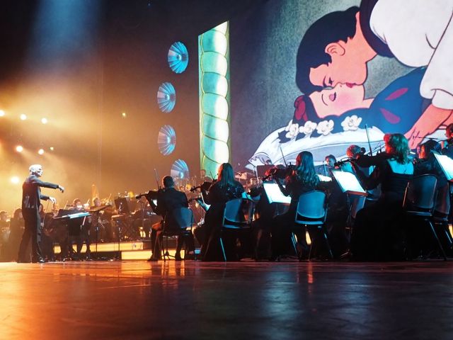 Das Bild zeigt Musiker auf der Bühne vor großen Bildschirmen, die Disney-Charaktere zeigen, darunter eines, das ein Paar beim Kuss aus dem Film Schneewittchen zeigt.