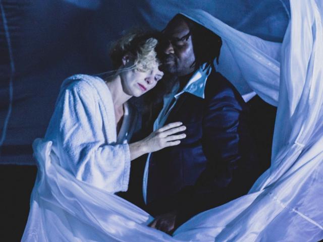 Das Bild zeigt eine Frau und einen Mann, die sich leidenschaftlich umarmen, vor dem Hintergrund eines schwarzen Vorhangs mit weißen Laken darauf, was an eine Szene aus der Theaterstück Othello erinnert.