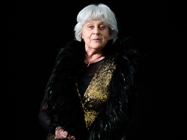 Das Bild zeigt eine ältere Frau mit einem gefiederten Boa, einem schwarzen Outfit mit einer goldenen Akzentweste und einem schwarzen Hut, die vor einem dunklen Hintergrund steht.
