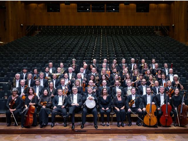 Eine Gruppe von Musikern, die bei einem Orchesterkonzert auftreten und alle in schwarzen Anzügen und Krawatten gekleidet sind