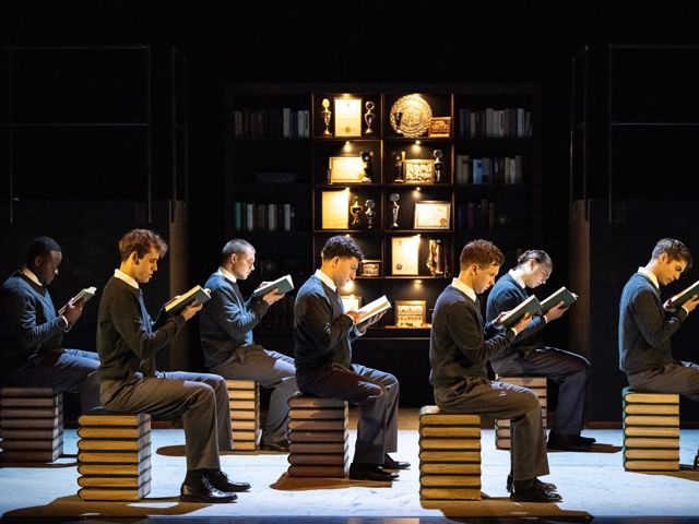 Eine Gruppe von Männern im Anzug sitzt auf Blöcken und liest Bücher, thereby creating a serious and reflective atmosphere.