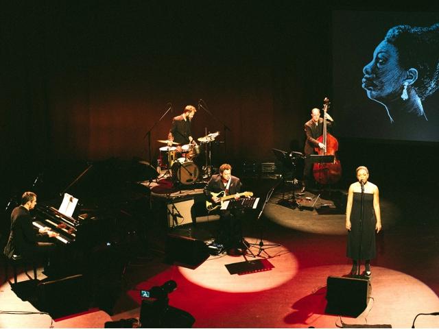 Person steht auf der Bühne mit einer Band, die hinter ihr spielt.