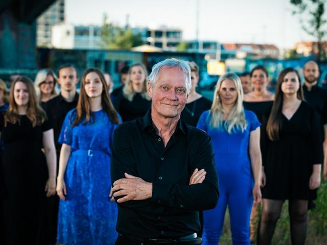 Alter Mann steht zwischen jungen Frauen in blauen Kleidern und lächelt in die Kamera.