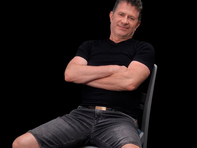 Ein Mann sitzt auf einem Stuhl, hat die Arme verschränkt und trägt ein schwarzes Hemd und Jeans-Shorts.
