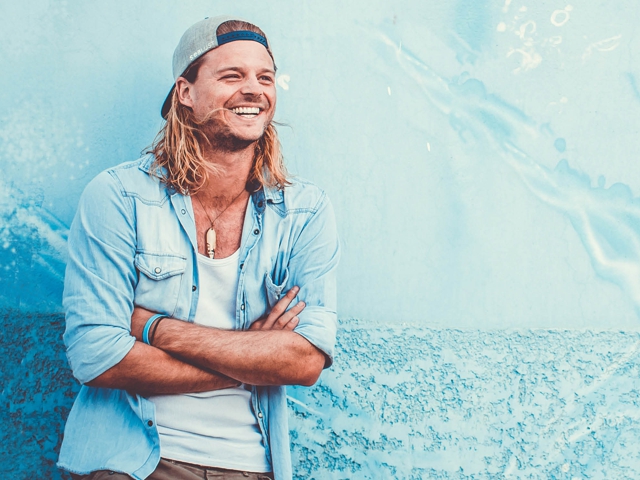 Der Mann hat langes Haar und steht vor einem Hintergrund, der vermuten lässt, dass er am Strand sein könnte, wie durch seine entspannte Pose und den wässrig-blauen Farbton angedeutet.