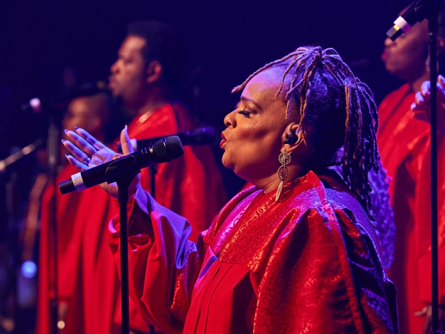 Der Sänger, der rote Roben trägt und vor einem Mikrofon steht, scheint Teil der Leistung der Harlem Gospel Singers zu sein.