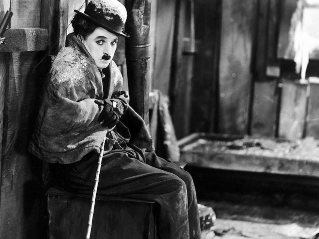 Ein Mann, der wie Charlie Chaplin gekleidet ist, sitzt vor Trümmern am Ende einer Eisenbahnstrecke.