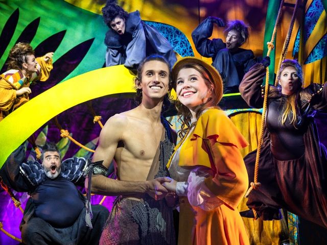 Das Bild zeigt eine Gruppe von Menschen auf der Bühne, darunter zwei zentrale Individuen, die offenbar die Hauptdarsteller einer Musicalaufführung sind und von anderen Performern in farbenfrohen Kostümen umgeben sind.