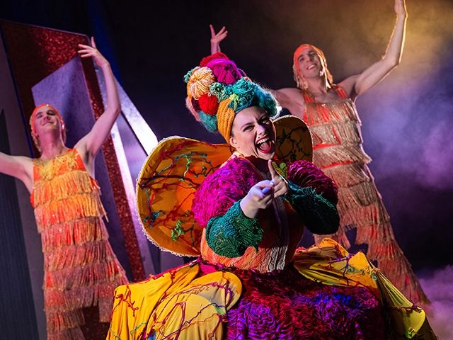 Frau als Dragqueen verkleidet auf der Bühne singend mit zwei Tänzern, die rechts und links von ihr tanzen