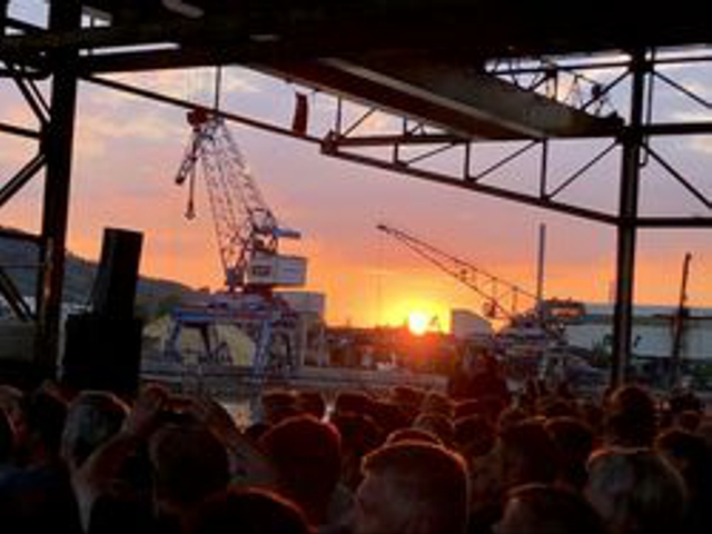 Das Foto zeigt eine Menge, die sich versammelt hat, um den Sonnenuntergang an einem industriellen Hafenbereich zu beobachten.