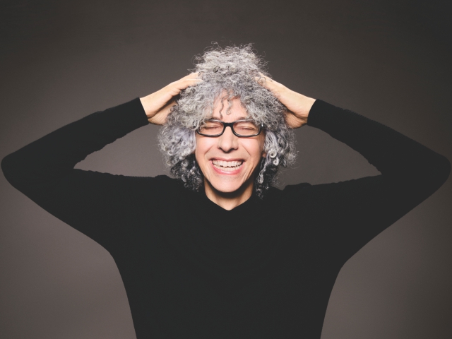 Eine Frau mit grauen Haaren und Brille, die lächelt und sich den Kopf berührt.
