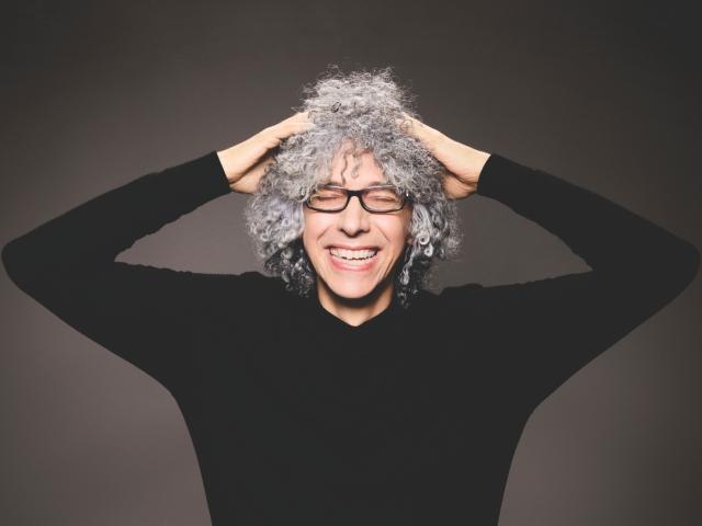 Eine Frau mit grauen Haaren und Brille, die lächelt und sich den Kopf berührt.
